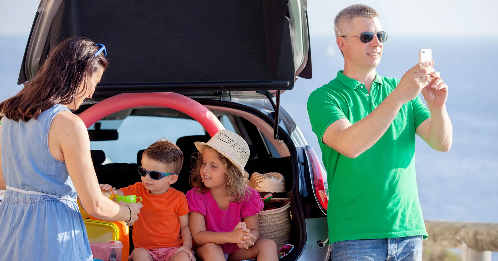 Een gezin gaat een dagje uit: kinder zitten in de kofferbak en krijgen wat te drinken van mama terwijl papa een foto aan het maken is van de omgeving